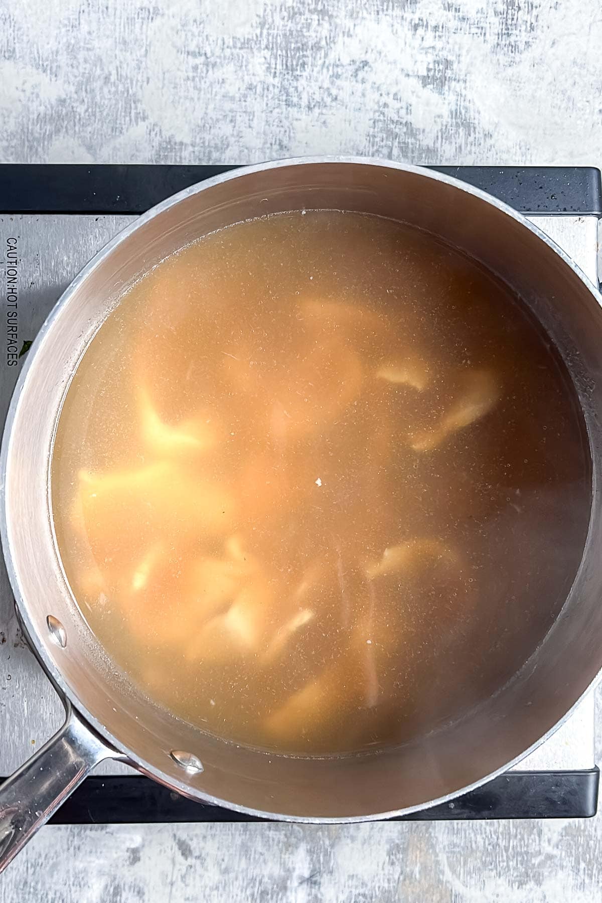 A metal pot filled with brown pho broth and thinly sliced chicken sits on a stovetop.