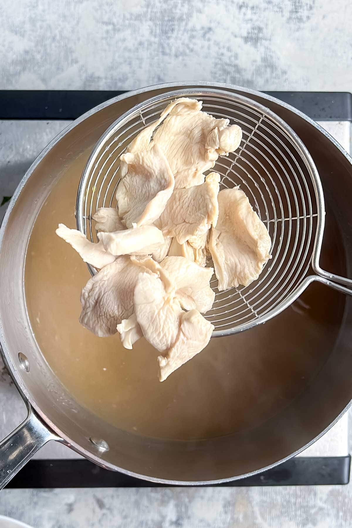 A slotted spoon lifts slices of cooked chicken breast from a pot of broth on a stovetop.