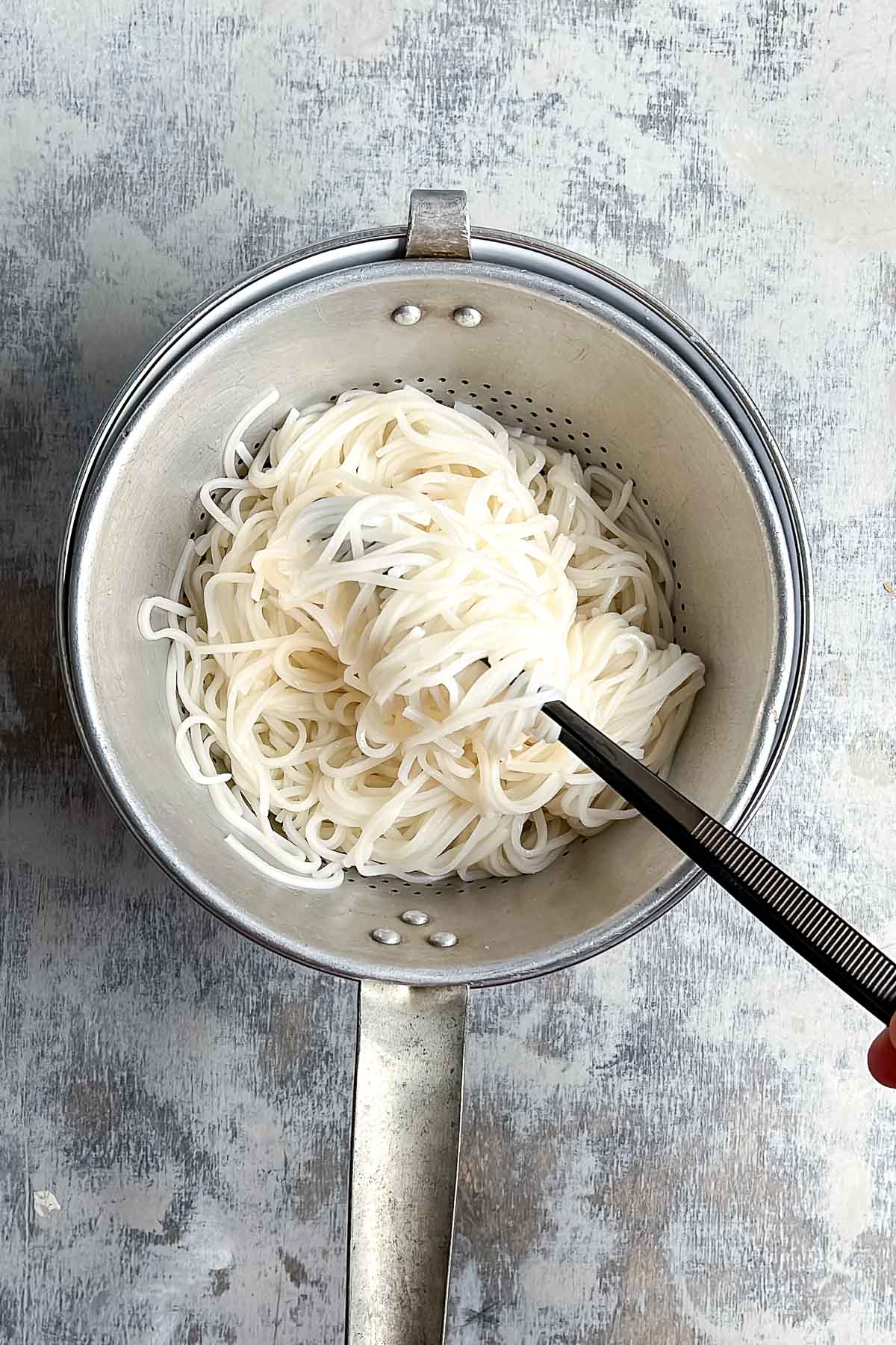 A metal colander filled with cooked white rice noodles, with tongs resting on top.