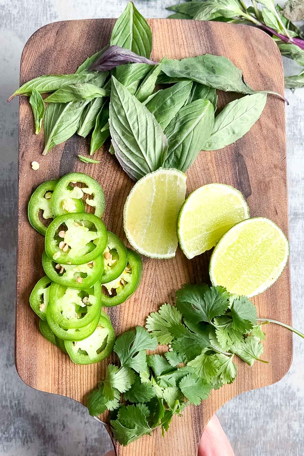 A wooden board with fresh basil leaves, cilantro, sliced jalapeño peppers, and two lime halves—perfect accompaniments for a steaming bowl of pho.