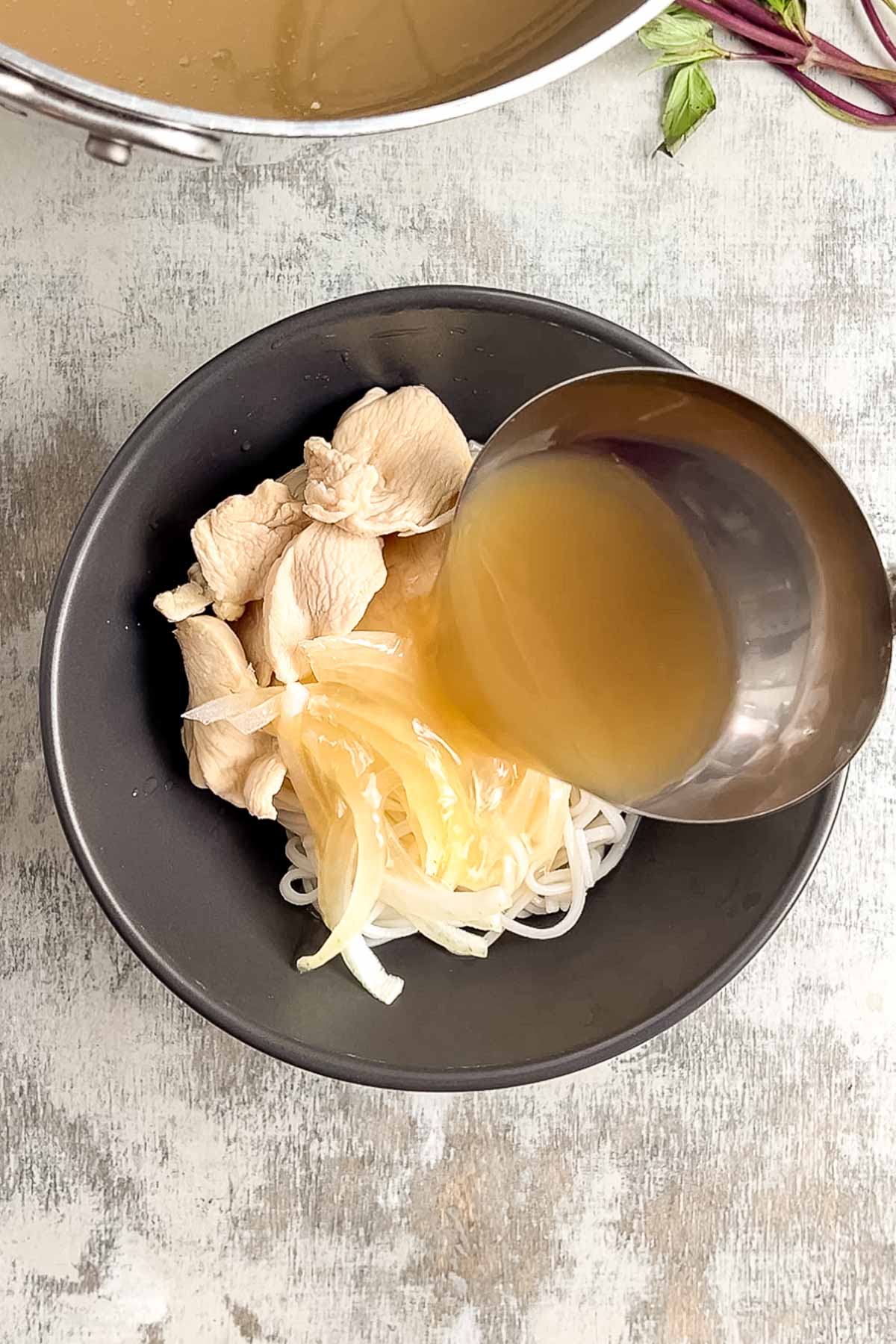 A metal ladle pours hot pho broth over sliced chicken, onions, and rice noodles in a black bowl on a textured surface.