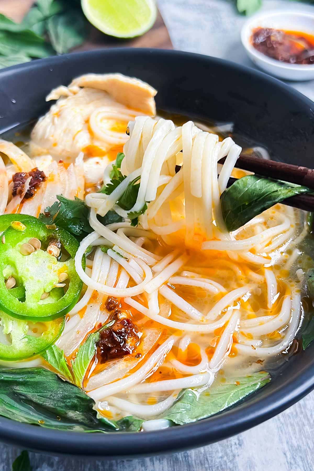 A bowl of pho noodle soup with slices of chicken, fresh herbs, jalapeño, and chili oil, with chopsticks lifting noodles.