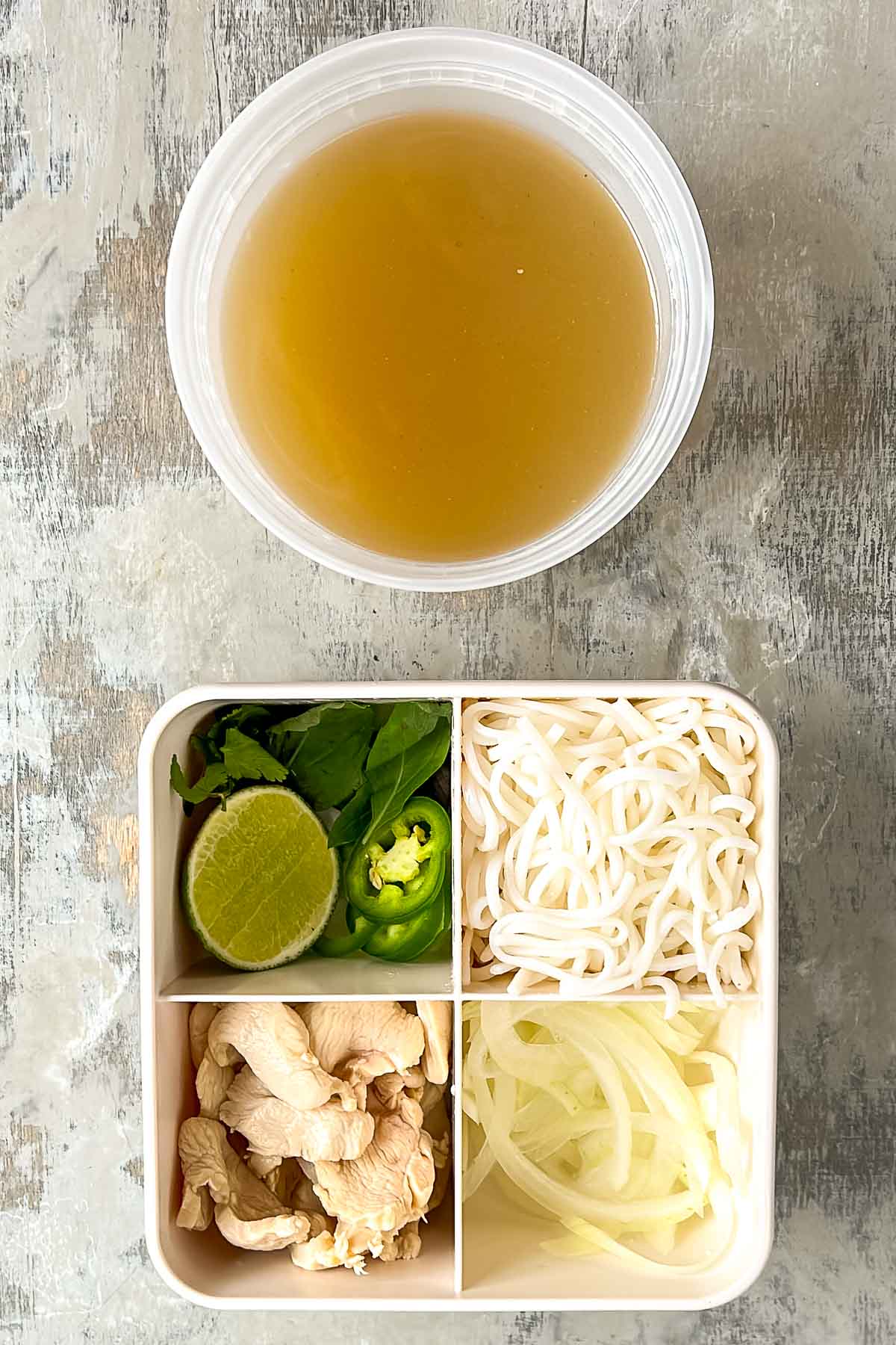 A container of pho broth sits above a divided tray with lime, herbs, jalapeño slices, rice noodles, sliced chicken, and onions on a light surface.