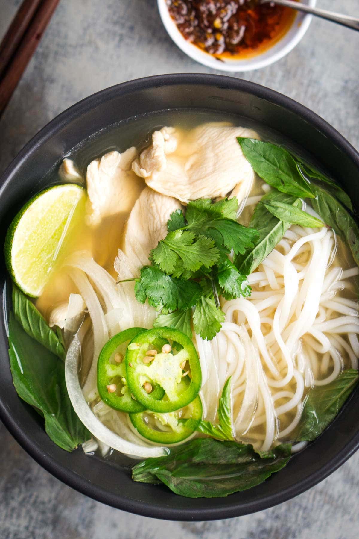 A bowl of pho with rice noodles, sliced chicken, fresh basil, cilantro, onion, and jalapeño slices next to a small bowl of chili oil.