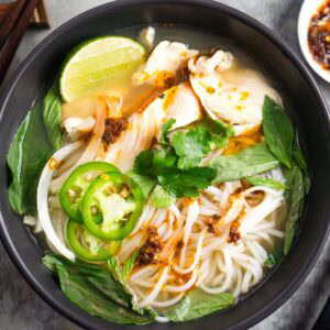 A bowl of chicken pho with rice noodles, sliced chicken, jalapeños, lime, cilantro, basil, onions, and chili oil in broth.