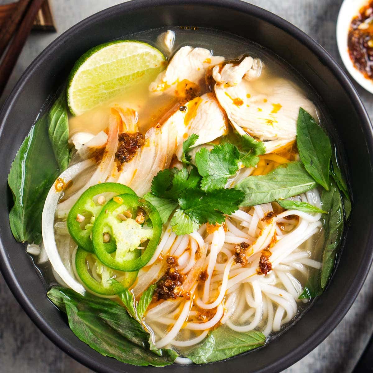 A bowl of chicken pho with rice noodles, sliced chicken, jalapeños, lime, cilantro, basil, onions, and chili oil in broth.