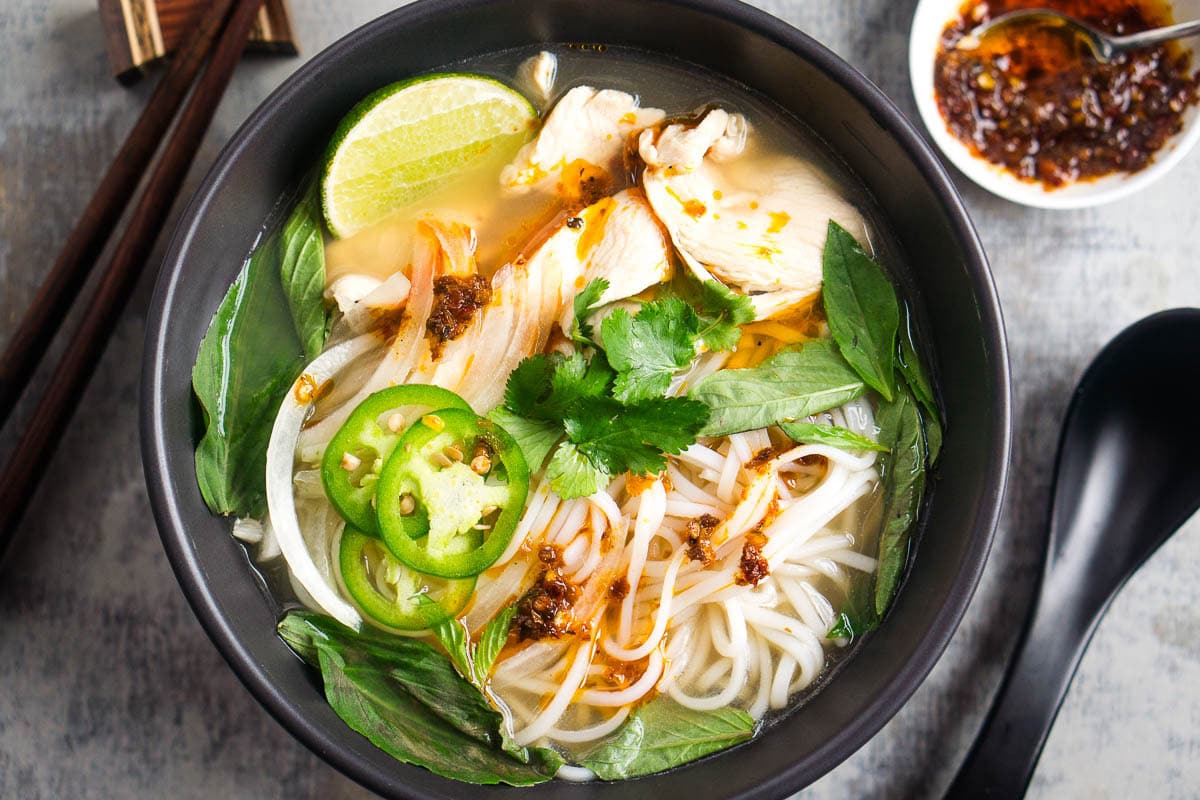 A bowl of chicken pho noodle soup with rice noodles, sliced jalapeños, lime, fresh herbs, and chili oil, with chopsticks, a spoon, and bowl of chili oil beside it.