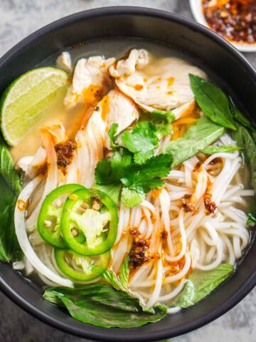 A bowl of pho noodle soup with slices of chicken, fresh herbs, jalape&ntilde;o, and chili oil.