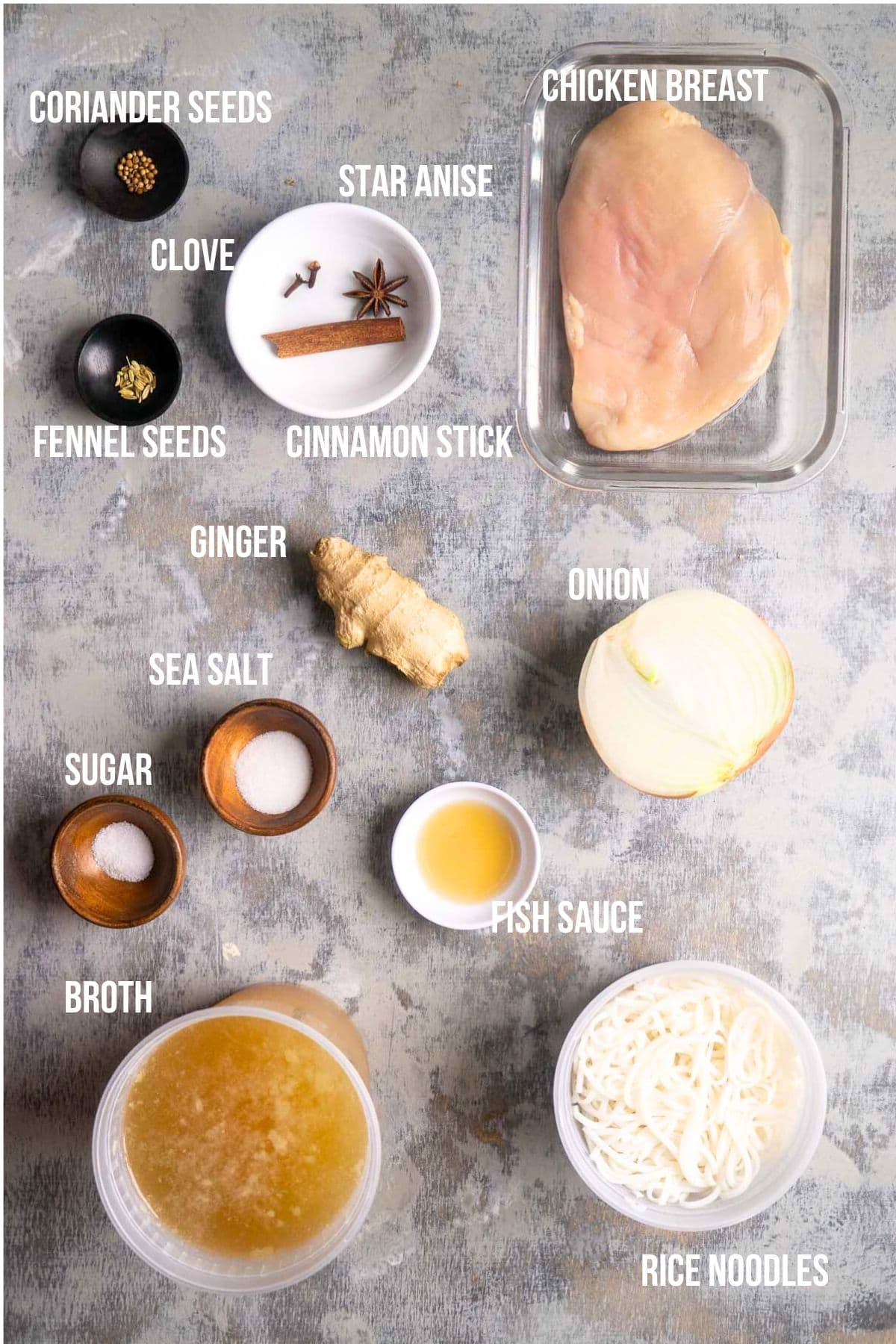 Assorted ingredients labeled for pho noodle soup including chicken breast, spices, onion, ginger, fish sauce, sea salt, sugar, broth, and rice noodles. All are arranged on a countertop in preparation for an easy pho soup recipe.