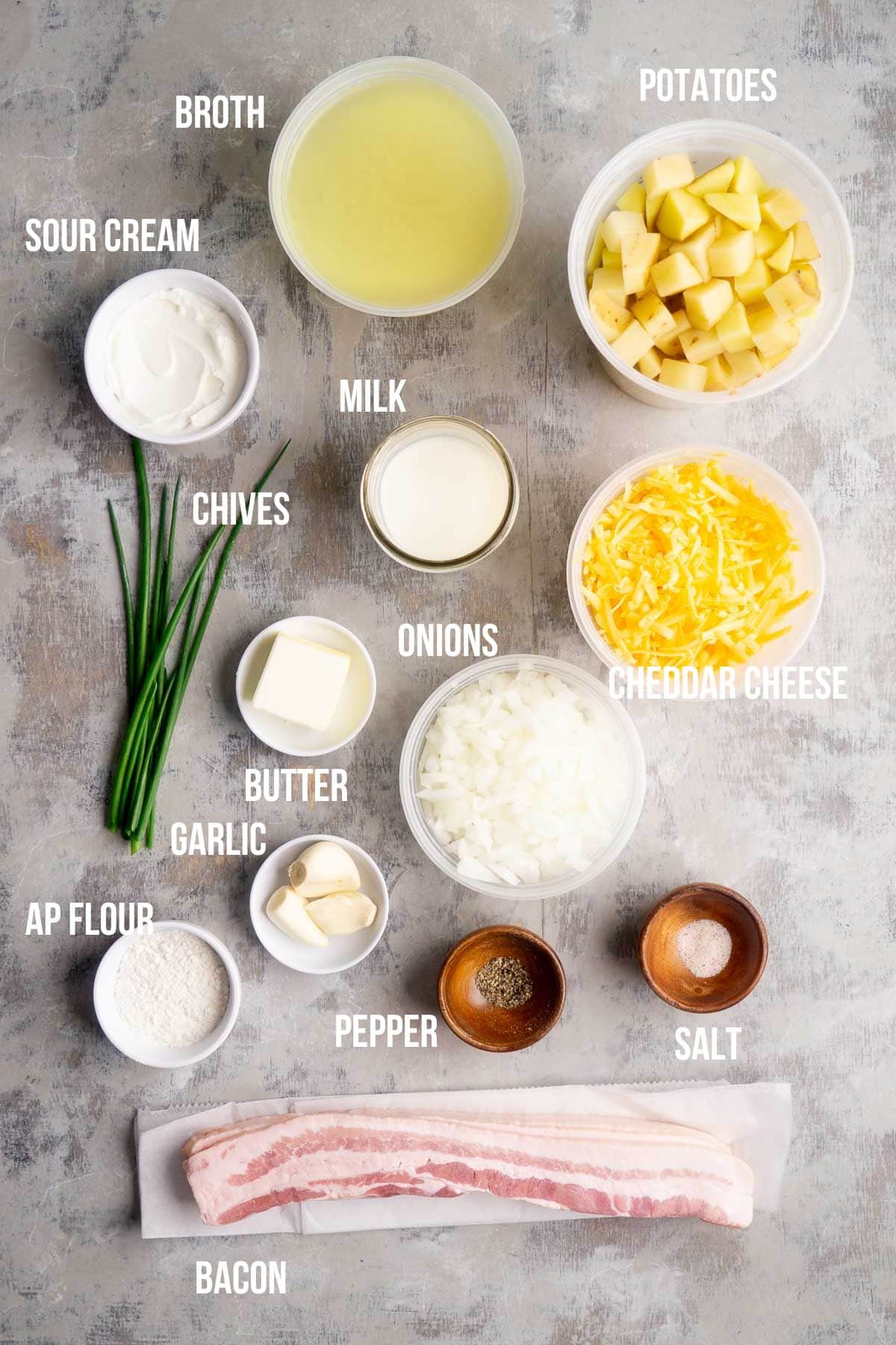 Ingredients for loaded potato soup arranged on a surface including broth, potatoes, sour cream, milk, chives, cheese, onions, butter, garlic, flour, pepper, salt, and bacon.