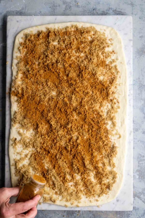 persons hand sprinkles cinnamon over rolled out dough with a layer of browns sugar
