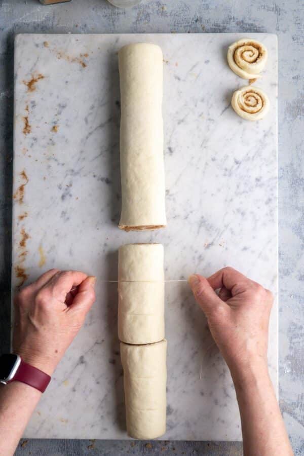 person's hands use dental floss to cut cinnamon roll dough log into pieces