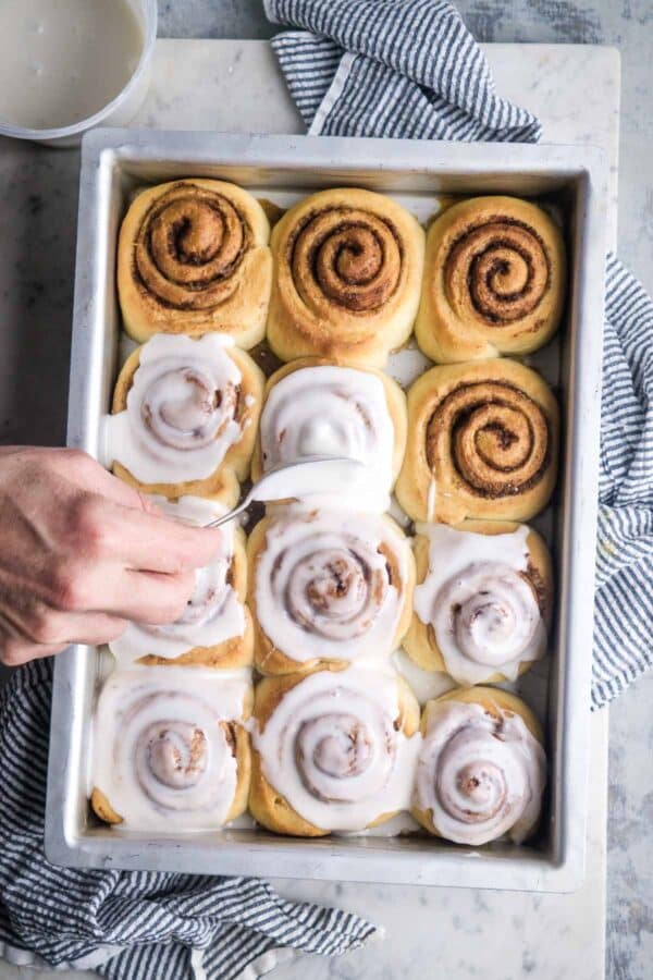 Persons hand applies glaze to cinnamon rolls with a spoon