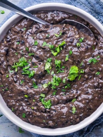 black refried beans garnished with fresh cilantro in white serving bowl next to lime wedges