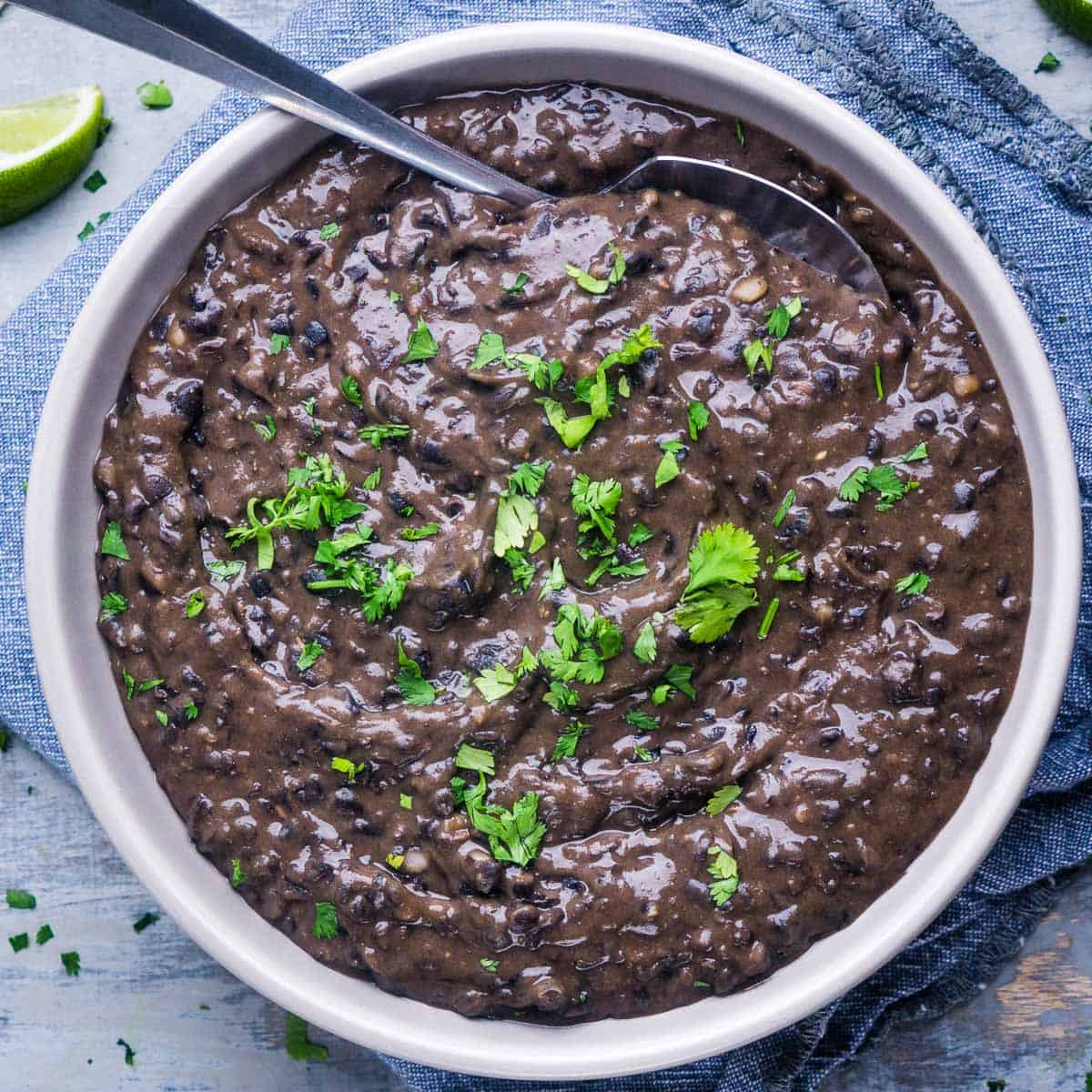 black refried beans garnished with fresh cilantro in white serving bowl next to lime wedges