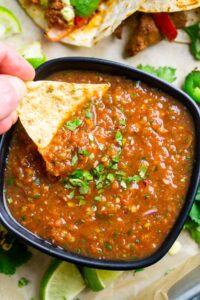 tortilla chip is dunked into blender salsa in a black bowl next to Mexican food