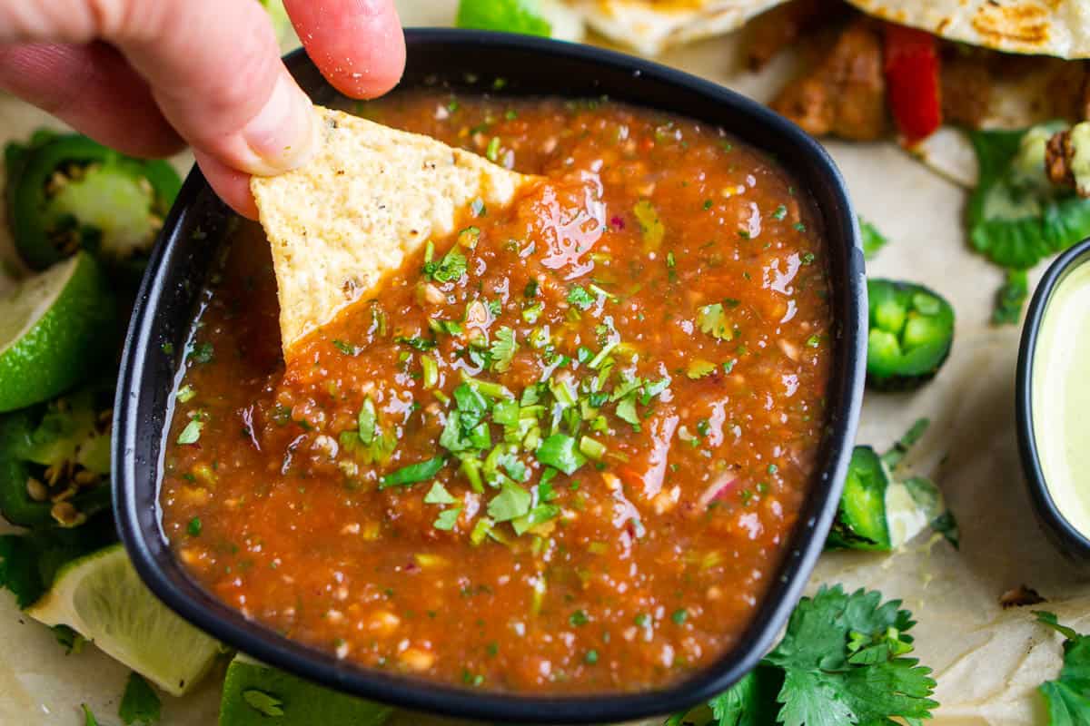 tortilla chip is dunked into blender salsa in a black bowl next to Mexican food