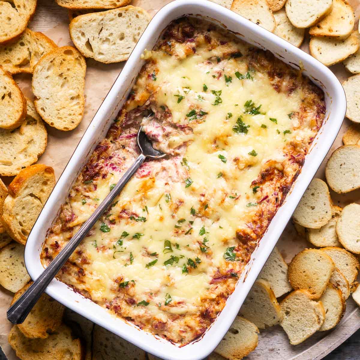 Rectangular dish filled with cheesy baked Reuben dip, garnished with chopped herbs and surrounded by toasted bread slices, with a spoon in the dish.