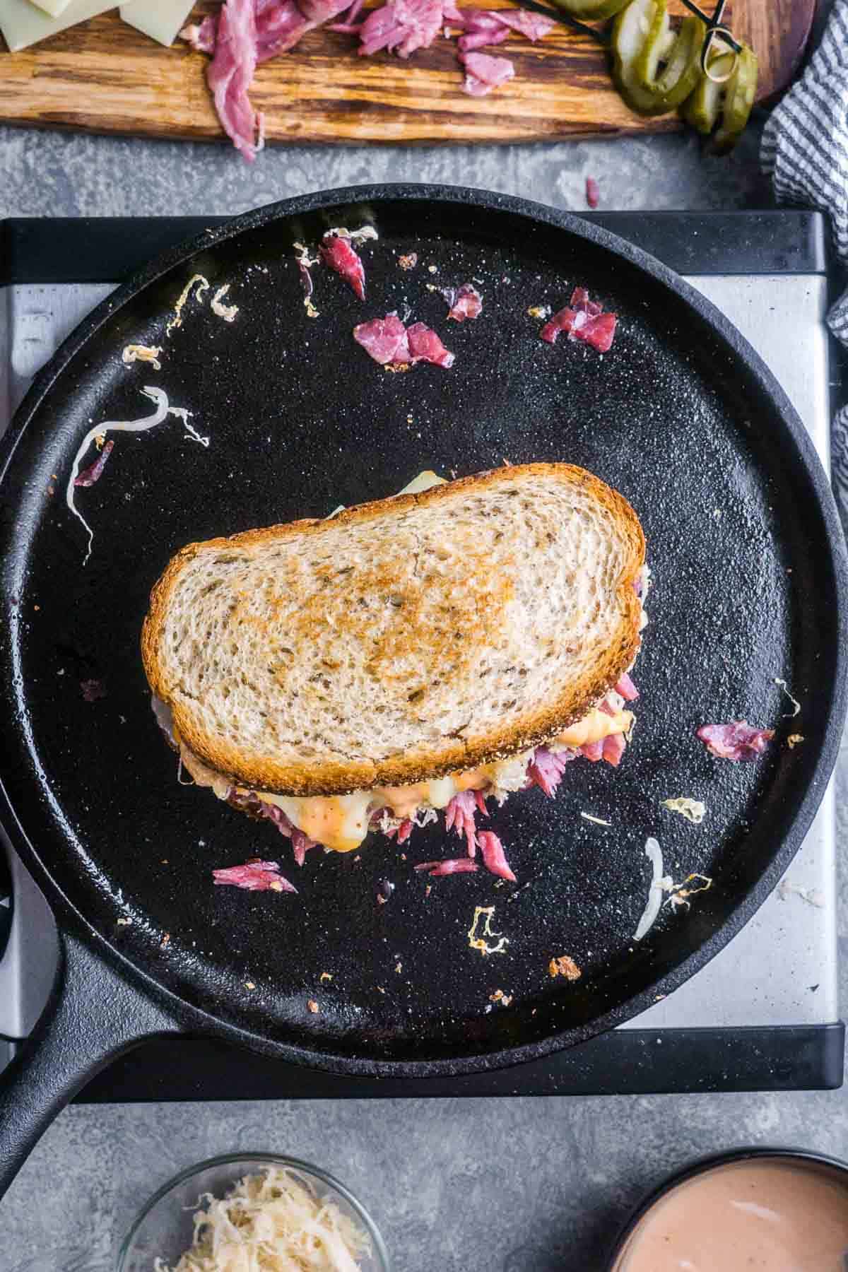 A corned beef Reuben sandwich is being grilled in a cast iron skillet, with some shredded cheese and extra corned beef scattered around.