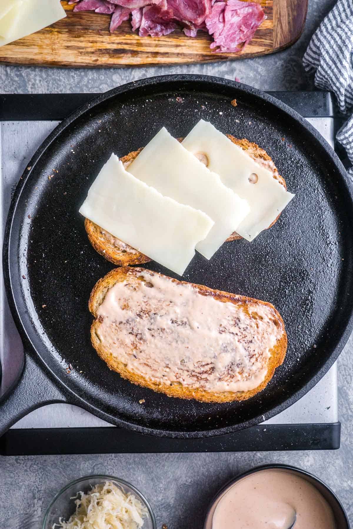 Two slices of rye in skillet; one topped with Russian dressing, the other topped with Swiss cheese