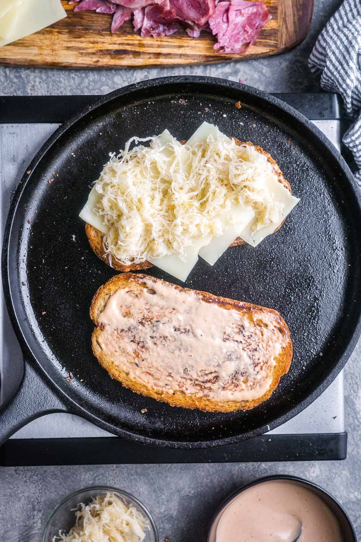 Two slices of rye bread in cast iron skillet; one topped with Russian dressing, the other topped with cheese and sauerkraut