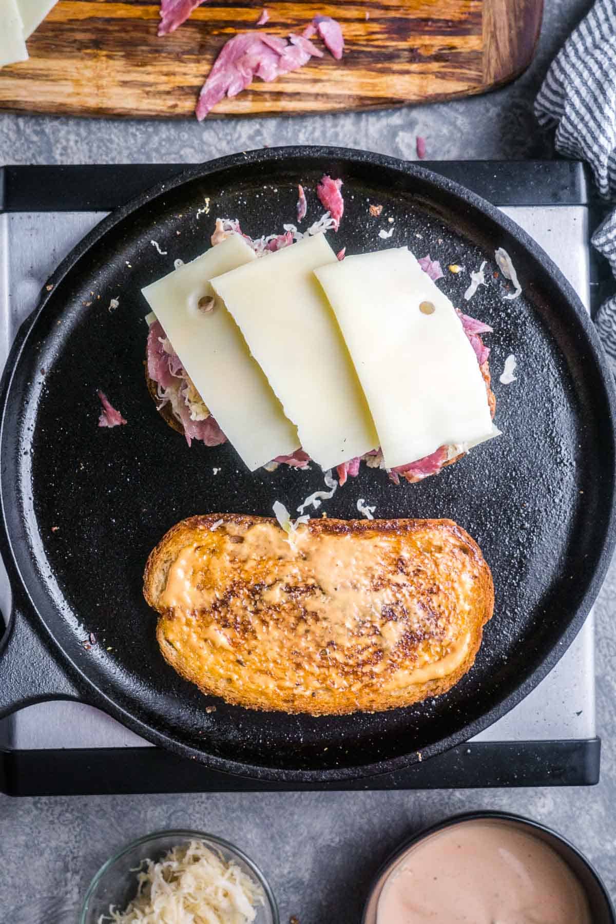 Overhead view of two Reubens in a cast iron skillet, one opened to show sliced cheese