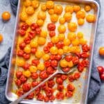 Red and yellow roasted cherry tomatoes and silver serving spoon on baking sheet with a blue linen underneath.