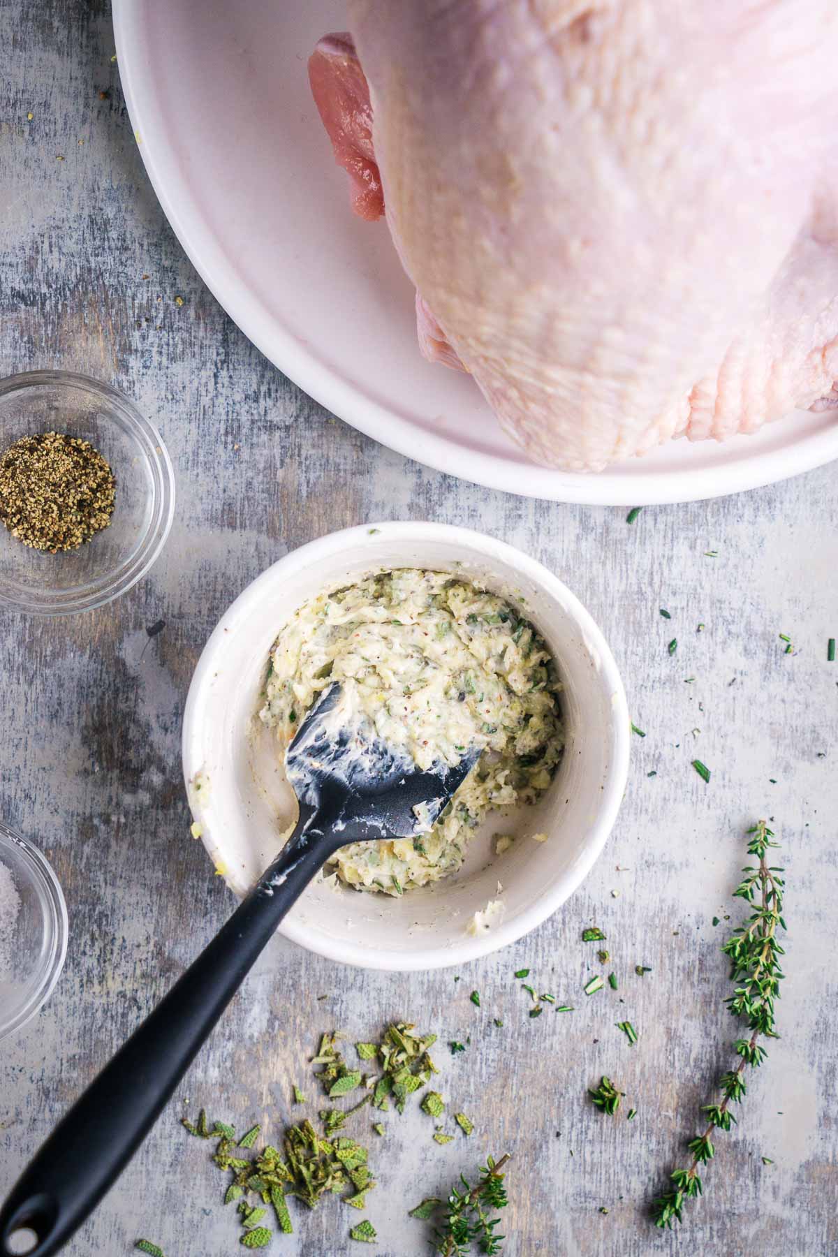 A white bowl containing herb butter mixed with a spatula, next to a raw whole turkey breast and sprigs of fresh herbs.