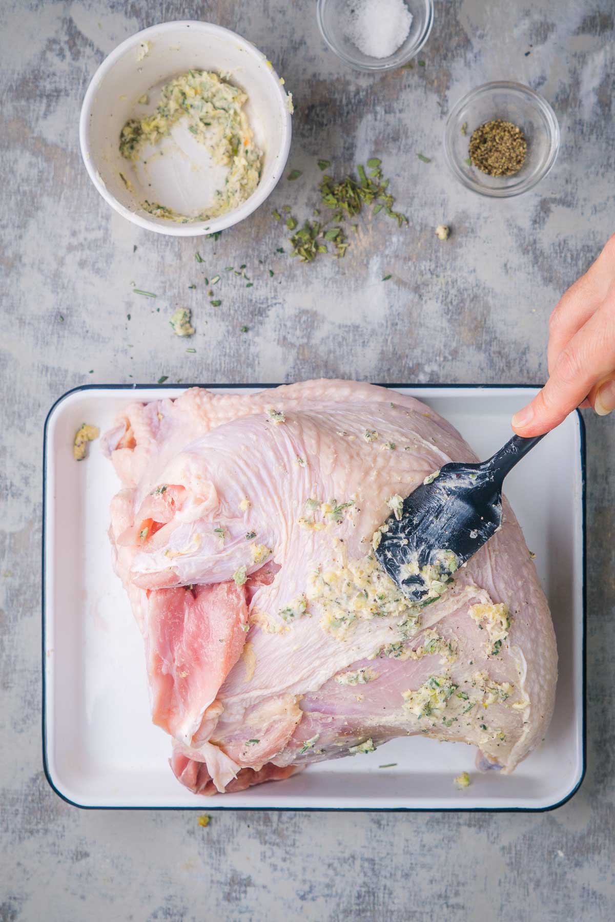 A hand uses a spatula to spread herb butter over a raw turkey breast in a baking pan.