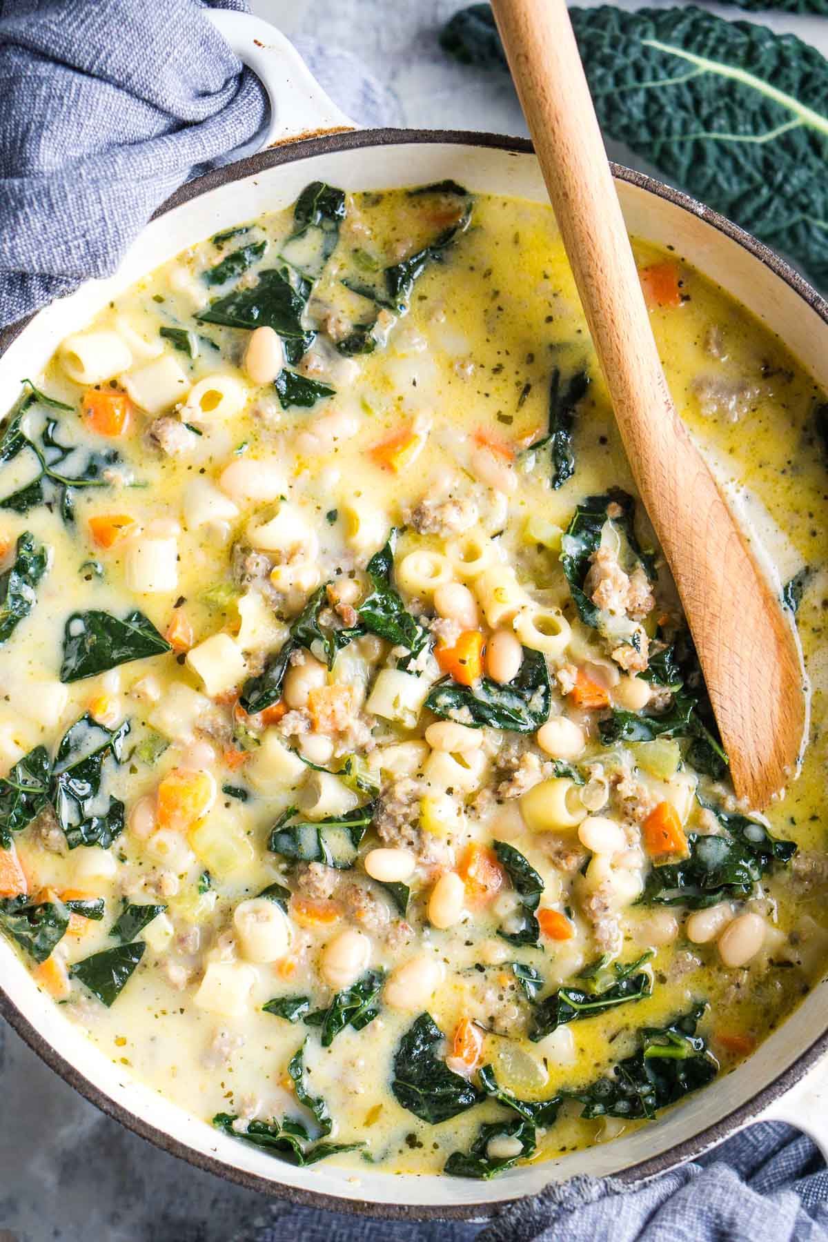 This might sound cliche, but one bite of this sausage, kale, and white bean soup will transport you to a cozy comfort zone, perfect for soup season. It’s quick, hearty, and satisfying, ideal for busy weeknights. With simple steps, even beginners can easily make it. The savory sausage, tender kale, creamy white beans, and rich broth create flavors that even picky eaters will love. It's become a family favorite at my house, and once you try it, I’m sure you’ll want it in your dinner rotation on repeat! Sausage and kale white bean soup in white soup pot with wooden soup ladle.
