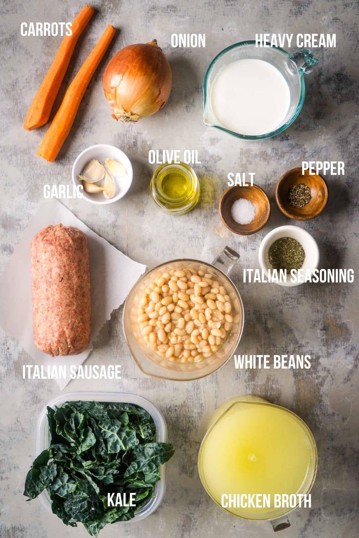 Ingredients for a recipe are arranged on a table, labeled: carrots, onion, heavy cream, garlic, olive oil, salt, pepper, Italian seasoning, Italian sausage, white beans, chicken broth, and kale.