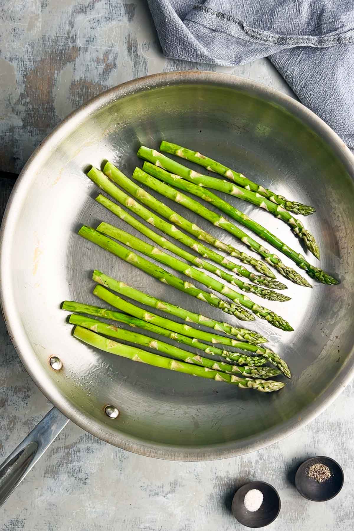 Asparagus spears are being sauteed in a stainless steel skillet, with small bowls of salt and pepper nearby on a textured surface.