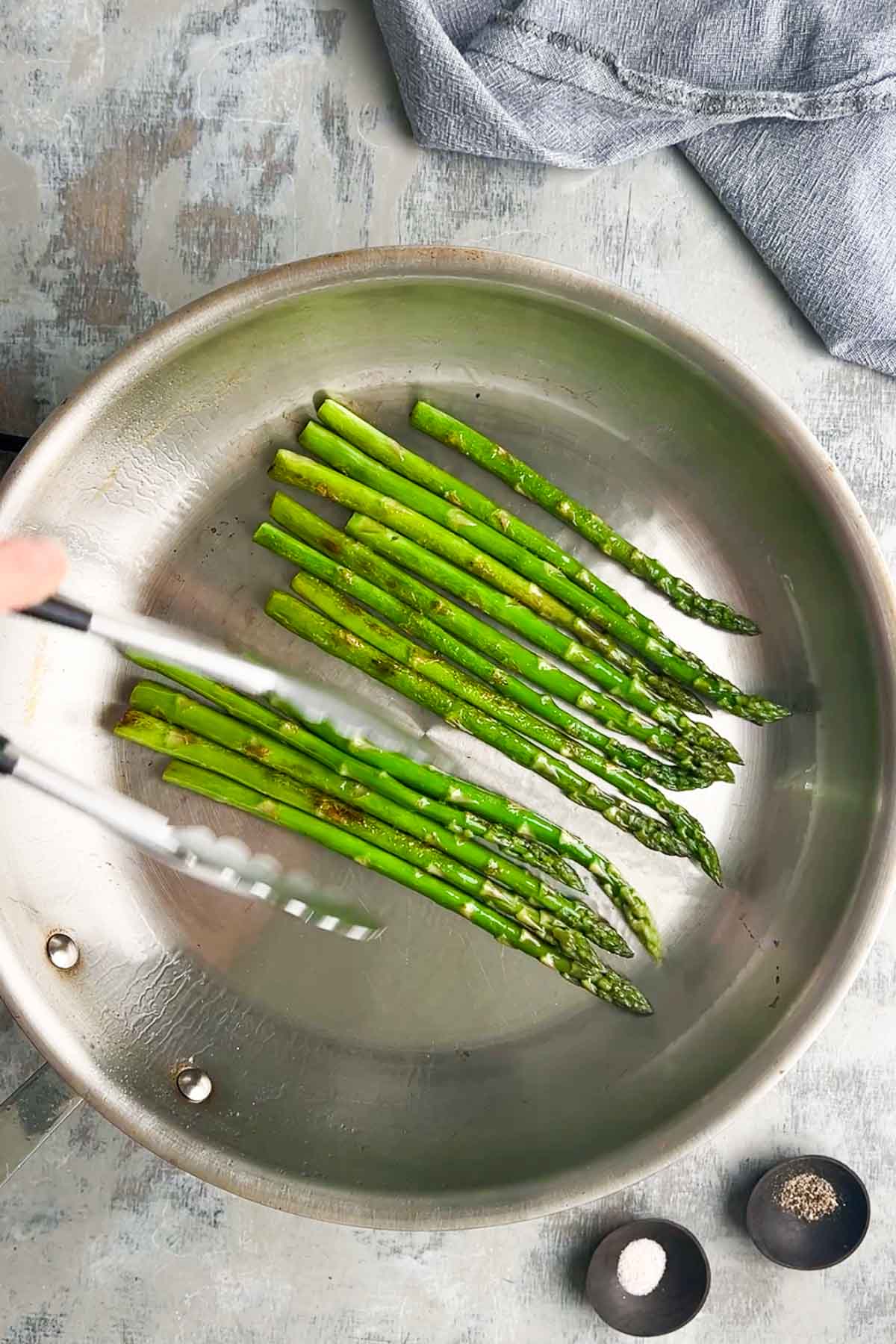 Asparagus spears are being sauteed in a stainless steel skillet, with small bowls of salt and pepper nearby on a textured surface.