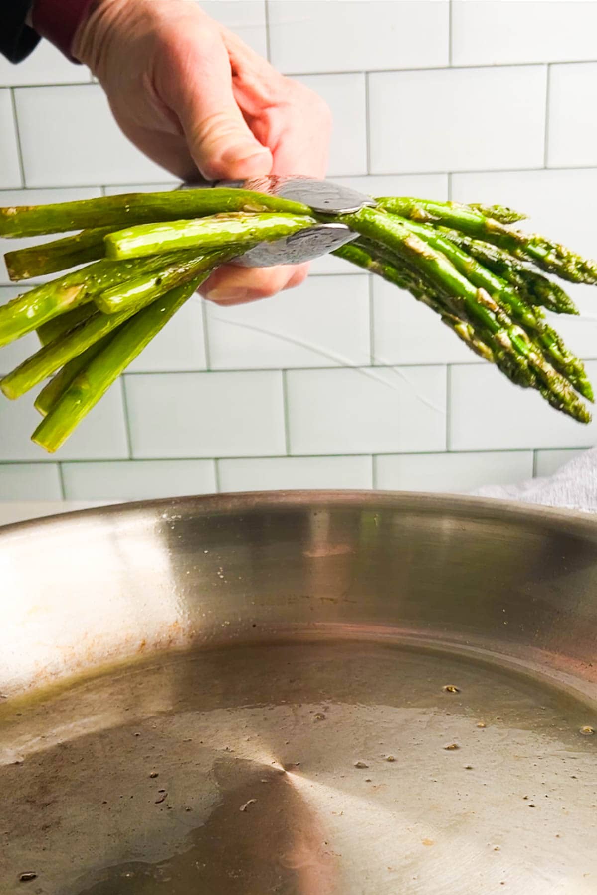 A hand holds a bunch of sauteed asparagus spears above a stainless steel pan, with a white tile backsplash in the background.