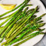Sauteed asparagus spears arranged on a white plate, garnished with lemon zest and served with a lemon wedge.