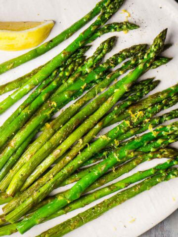 Sauteed asparagus spears arranged on a white plate, garnished with lemon zest and served with a lemon wedge.