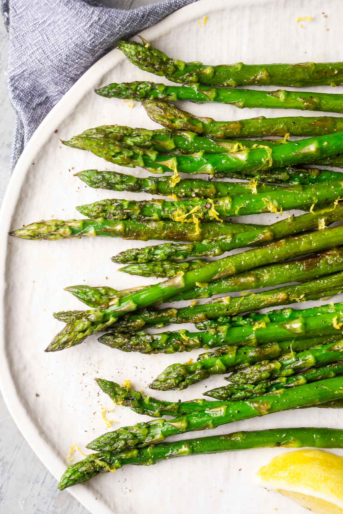 Sauteed asparagus spears arranged on a white plate, garnished with lemon zest and served with a lemon wedge.