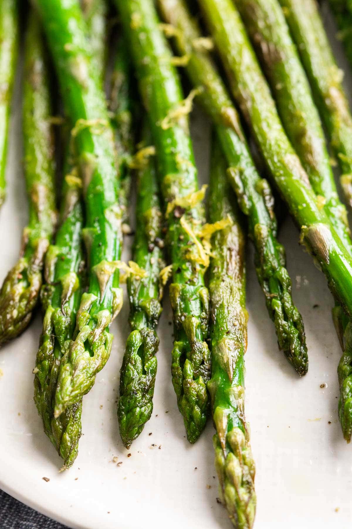 Sauteed asparagus spears arranged on a white plate, garnished with lemon zest and served with a lemon wedge.