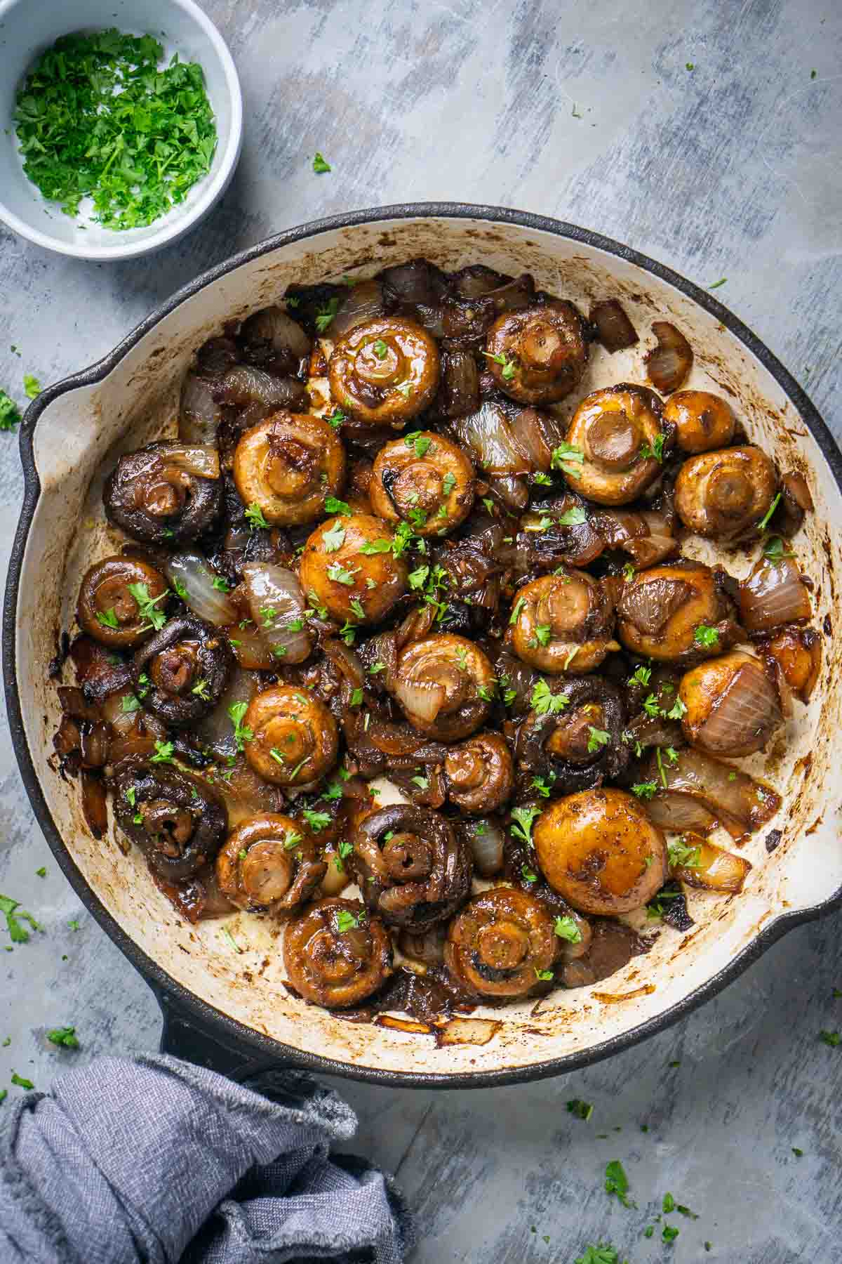 Cast iron skillet with cooked mushrooms and onions, garnished with chopped parsley. A small bowl with more parsley is in the background.