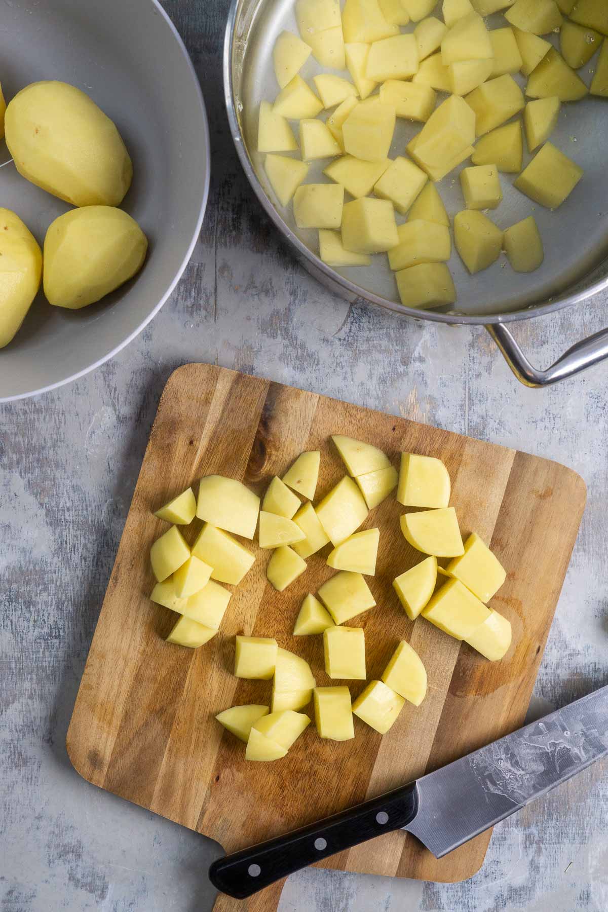 chef knife and chopped potatoes on cutting board next to stainless pot with diced potatoes next to peeled whole potatoes in bowl