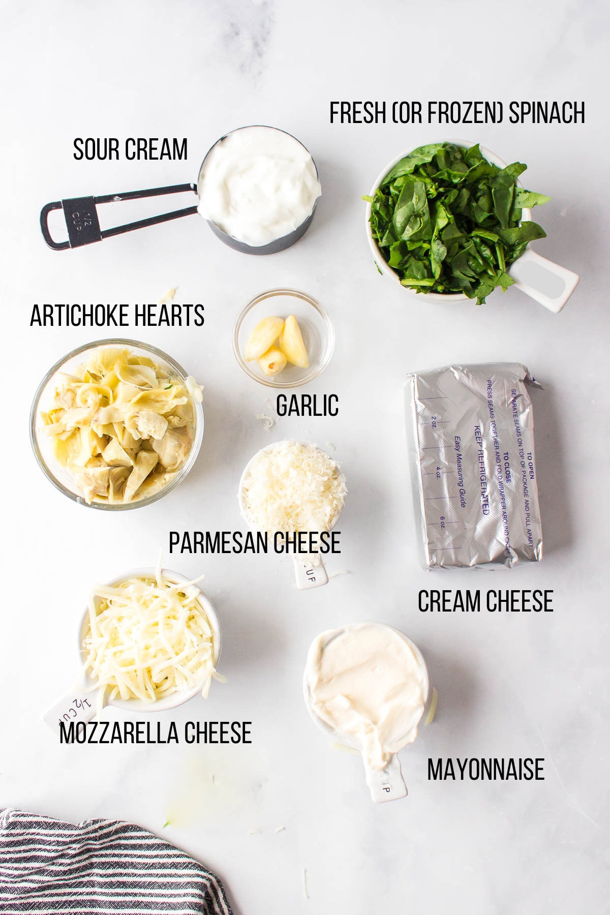 Ingredients on a white surface for spinach artichoke dip: sour cream, artichoke hearts, garlic, parmesan cheese, mozzarella cheese, mayonnaise, cream cheese, and fresh (or frozen) spinach—each clearly labeled.