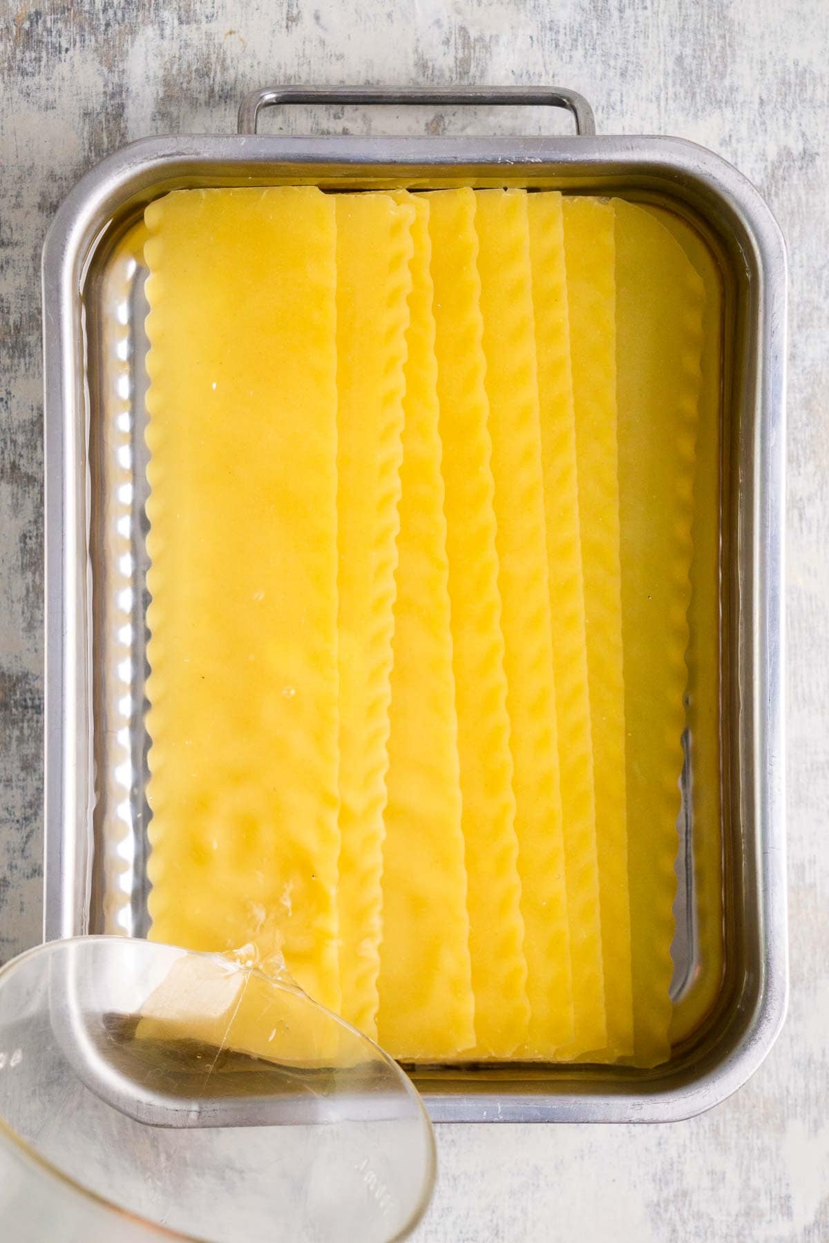 Uncooked lasagna noodles are arranged in a metal baking dish with water being poured over them from a glass container.