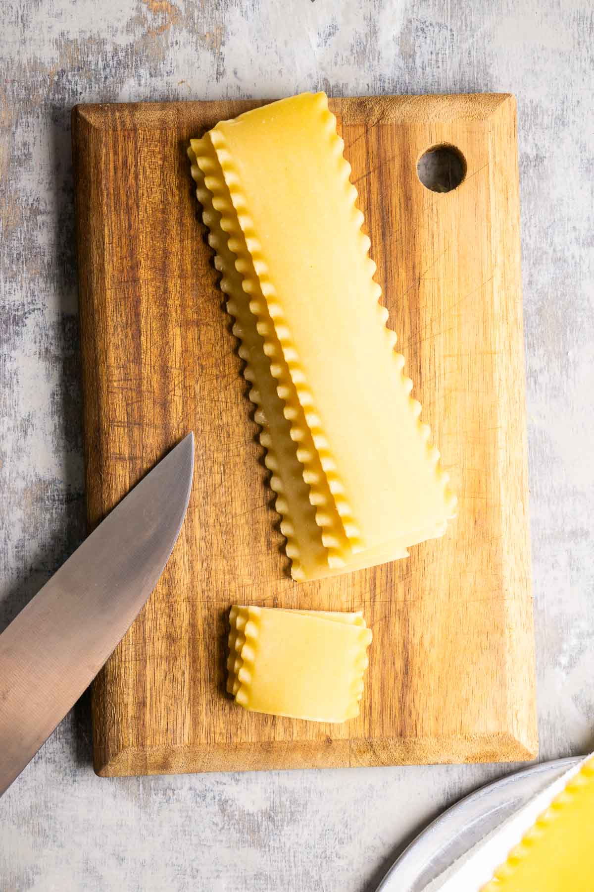 A stack of uncooked lasagna noodles, one cut into a small square, sits on a wooden cutting board next to a large kitchen knife.