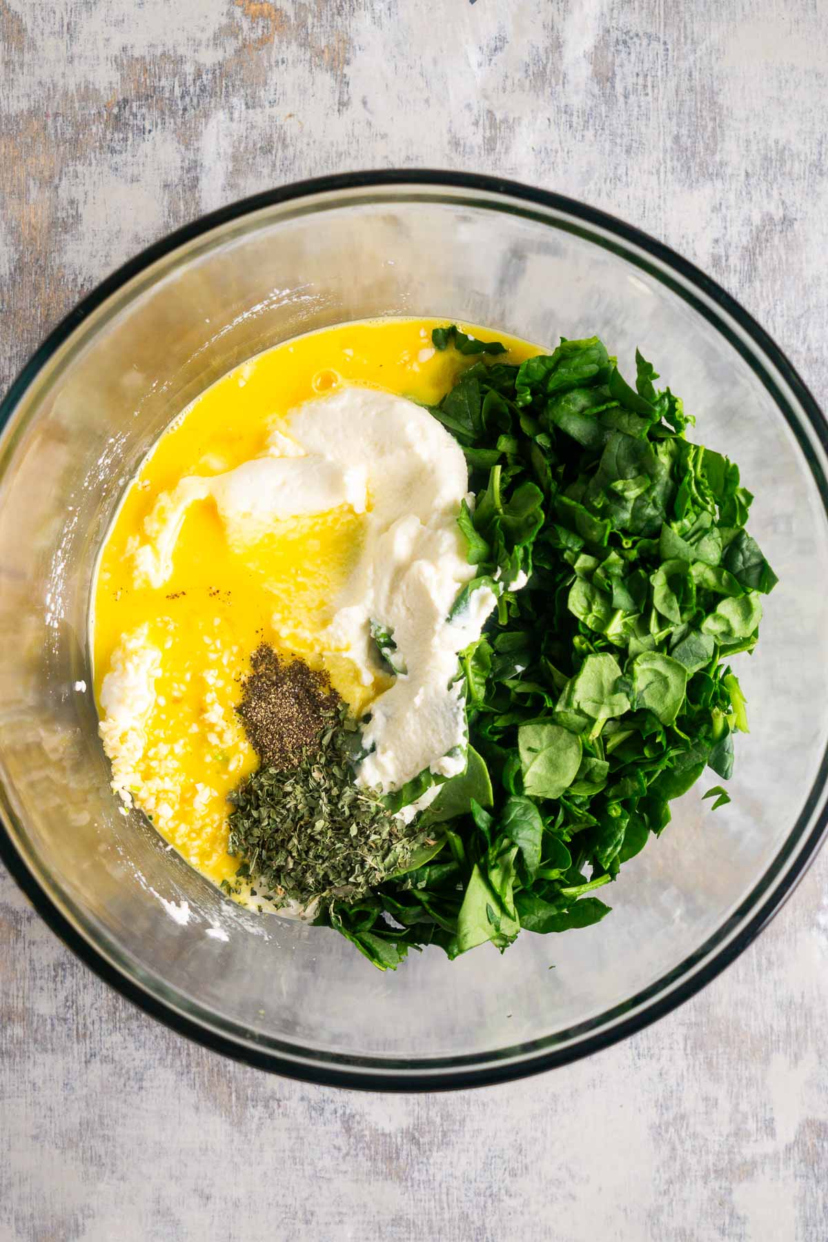 Top down view of a glass bowl containing beaten eggs, chopped fresh spinach, ricotta cheese, dried herbs, black pepper, and salt&mdash;classic ingredients for spinach ricotta lasagna.
