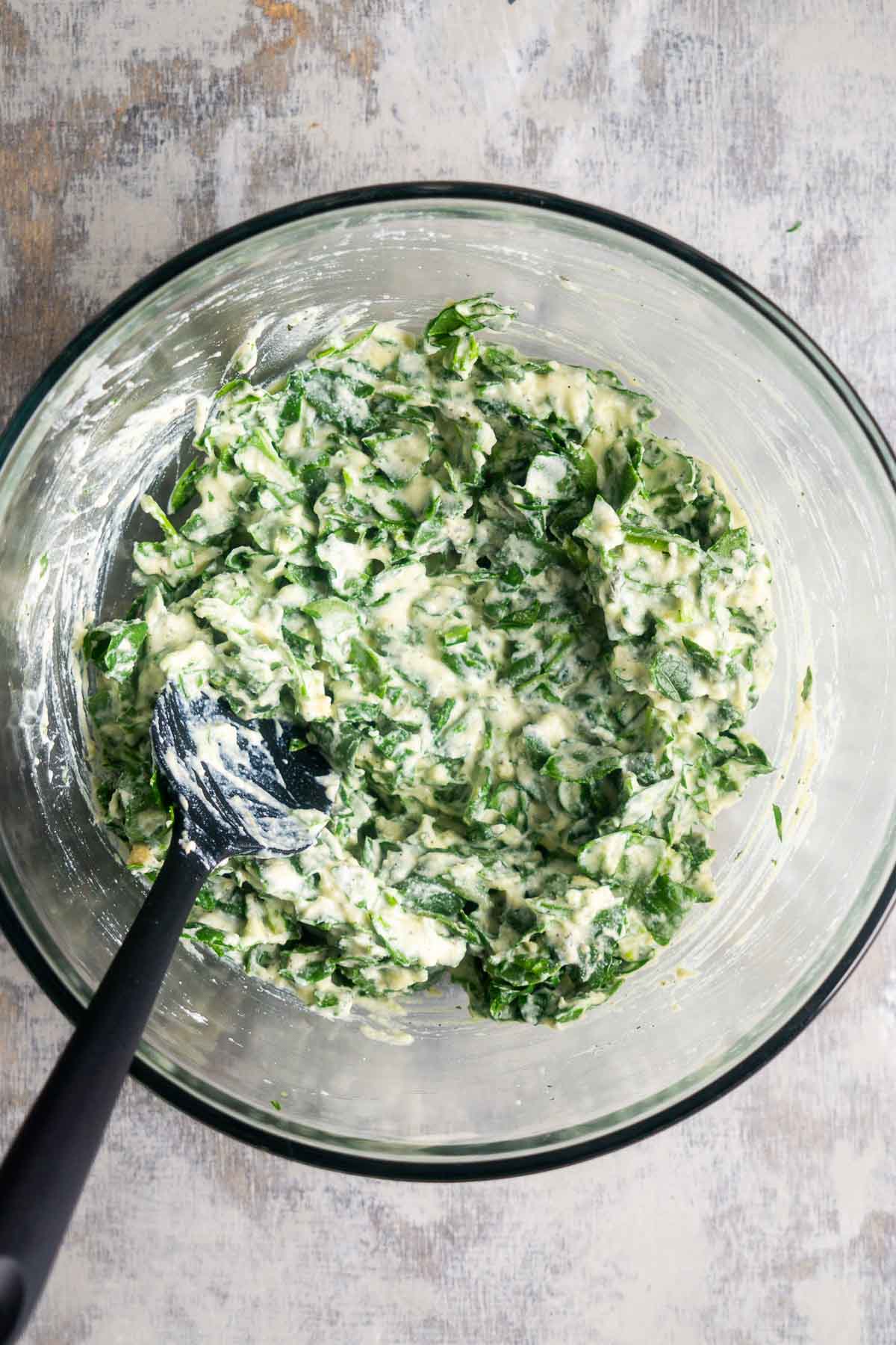 A glass bowl containing a creamy mixture of chopped fresh spinach and ricotta cheese is being mixed with a black spatula.