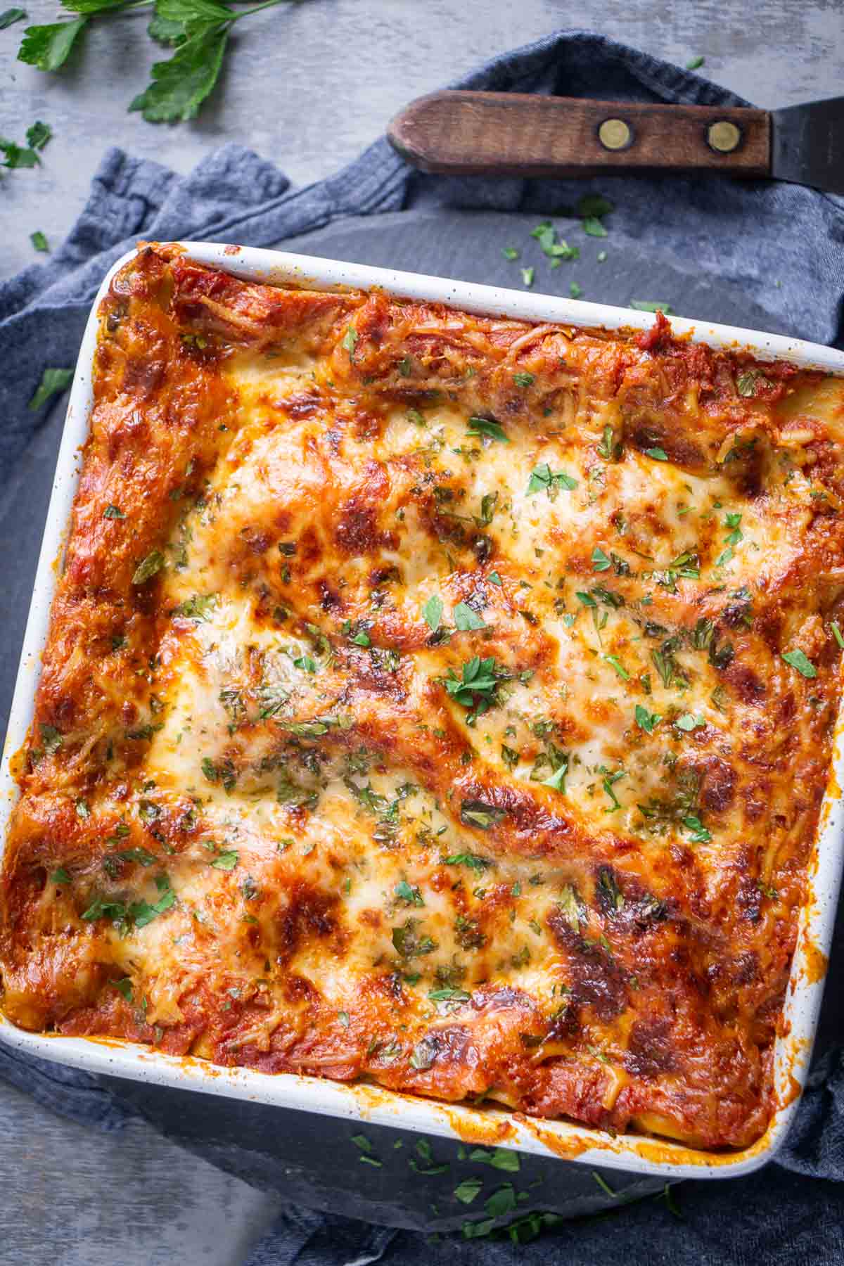 A baked spinach lasagna in a white dish topped with melted cheese and sprinkled with chopped herbs sits on a dark cloth next to a wooden-handled spatula.