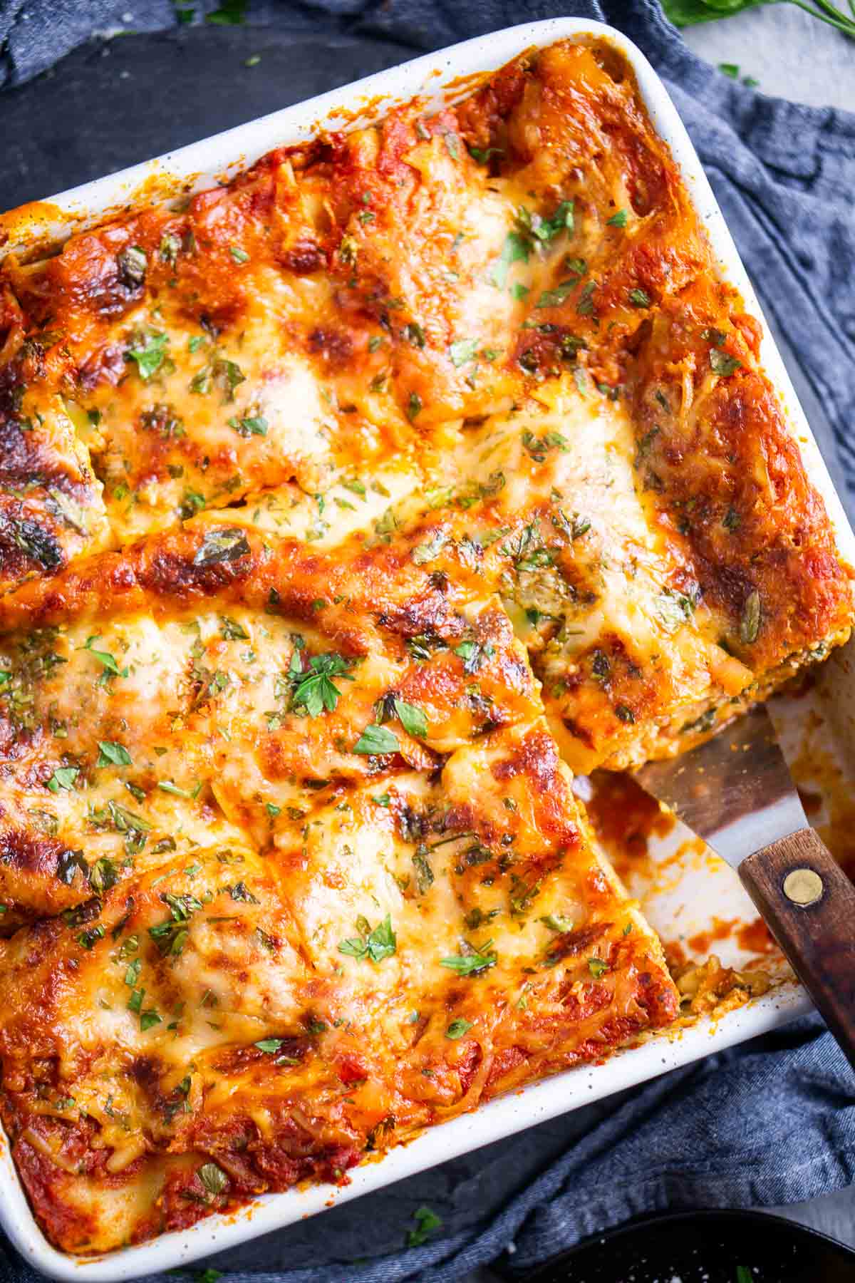 A rectangular baking dish filled with baked spinach lasagna, topped with melted cheese and herbs; one slice is being lifted out with a spatula.