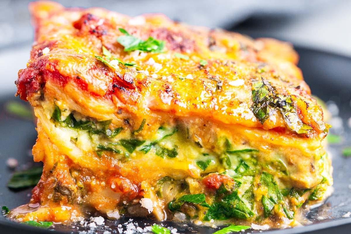 A close-up of a slice of spinach and ricotta lasagna, featuring layers of spinach, melted cheese, tomato sauce, and herbs, served on a dark plate.