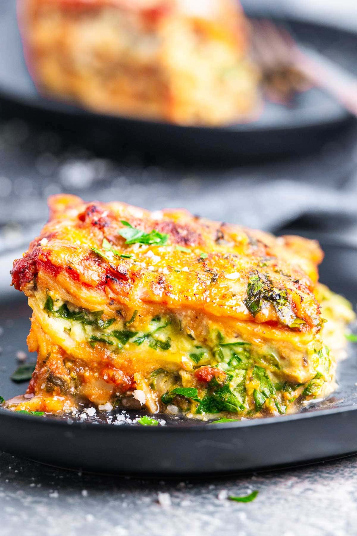 A close-up of a slice of spinach and ricotta lasagna, featuring layers of spinach, melted cheese, tomato sauce, and herbs, served on a dark plate.