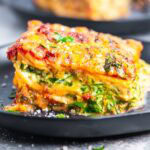 A close-up of a slice of spinach and ricotta lasagna, featuring layers of spinach, melted cheese, tomato sauce, and herbs, served on a dark plate.